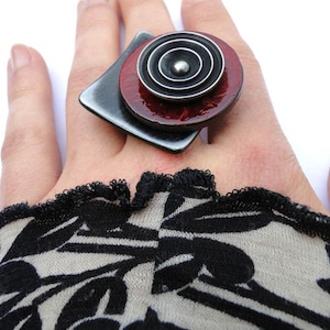 May include: A statement ring with a geometric design. It features a dark grey square base, a circular red element, and a silver spiral. The ring is a bold piece of jewellery.