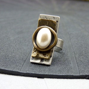 May include: A silver and brass ring with a white pearl set in a textured brass frame. The ring is on a grey surface.