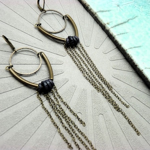Peut inclure: Une paire de boucles d'oreilles dorées avec un cercle et une barre incurvée. Chaque boucle d'oreille a un groupe de petites perles noires et plusieurs chaînes pendantes.