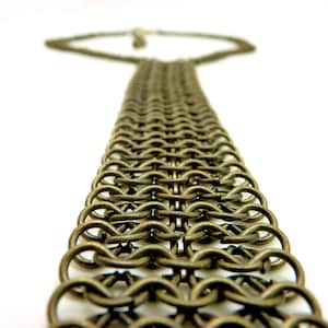 May include: A necklace featuring a wide, flat band constructed from interlocking bronze-toned metal rings in a chainmail pattern. The band extends into a thinner chain that connects at the back, showcasing a uniform, repetitive circular link design.