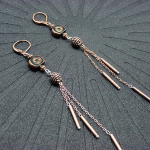 Puede incluir: Un par de pendientes colgantes con cuentas cuadradas negras y verdes, cadena de cobre y cuentas de cobre y plata. Los pendientes tienen un diseño de borla con múltiples hebras de cadena y varillas de cobre.