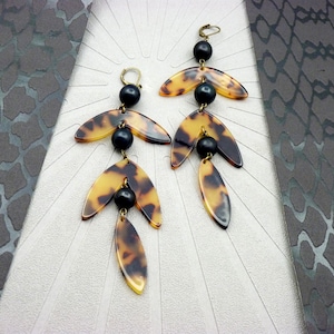 Peut inclure: Une paire de boucles d'oreilles pendantes avec un motif écaille de tortue. Chaque boucle d'oreille comporte trois éléments en forme de feuille, reliés par des perles noires et des cerceaux dorés. Les boucles sont présentées sur une surface blanche.