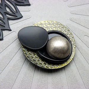 Peut inclure: Objet décoratif en métal avec une forme ovale noire et une sphère argentée sur une base dorée et noire à motifs. L'objet est posé sur une surface grise texturée, avec un élément décoratif noir en haut à gauche.