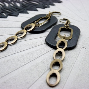 Op de afbeelding: Een paar oorhangers met een donkergrijze rechthoekige basis en messingkleurige metalen details. Elke oorbel heeft een reeks ovaalvormige schakels in messingkleur die aan de basis hangen. Gepresenteerd op een getextureerd, lichtgrijs oppervlak.