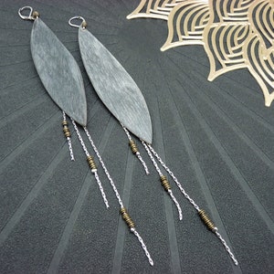 Peut inclure: Une paire de longues boucles d'oreilles en bois gris avec des chaînes argentées et des perles bronze. Les boucles d'oreilles sont suspendues à une surface gris foncé.