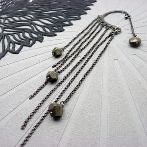 Könnte beinhalten: Ein silberfarbener Ohrschmuck mit herabhängenden Ketten und facettierten Pyritperlen. Der Ohrschmuck hat ein gebogenes Design, das sich um das Ohr legt, mit mehreren Ketten unterschiedlicher Länge. Die Ketten sind mit kleinen, geometrischen Pyritperlen verziert.