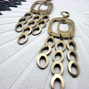 Peut inclure: Une paire de boucles d'oreilles de couleur laiton. Chaque boucle d'oreille présente un dessus carré avec un carré plus petit à l'intérieur, et trois chaînes pendantes de formes ovales. Les boucles d'oreilles sont présentées sur une surface texturée gris clair.