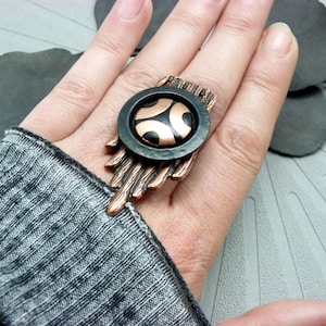 Peut inclure: Une bague composée d'un disque central noir orné d'un motif géométrique cuivré, montée sur une base en cuivre texturée rappelant des branches qui s'étend verticalement sur le doigt, créant un contraste entre les tons sombres et métalliques.