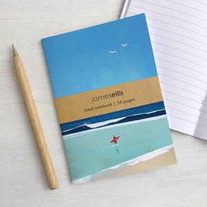 Puede incluir: Un cuaderno de rayas con una ilustración de portada de un surfista sobre una tabla roja en agua turquesa bajo un cielo azul con dos aves blancas. Una banda de papel color canela dice jamesellis lined notebook | 36 pages. Un bolígrafo de madera.