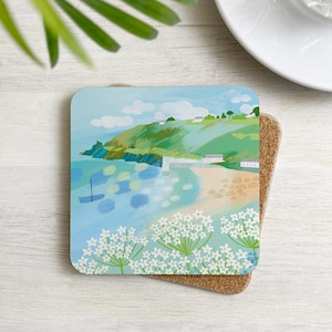 Puede incluir: Posavasos cuadrado con una ilustración de paisaje costero. El diseño presenta una playa, colinas verdes, agua azul y flores blancas. El posavasos está colocado sobre una base de corcho.