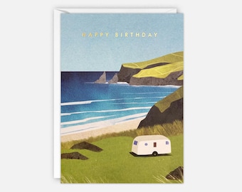 Op de afbeelding: Een verjaardagskaart met een witte caravan geparkeerd op een grassige heuvel met uitzicht op een strand met blauw water en groene kliffen. De tekst "HAPPY BIRTHDAY" is in goud gedrukt bovenaan de kaart.