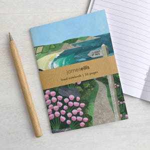 Op de afbeelding: Een gelinieerd notitieboek met een omslagillustratie van een kustlandschap, een pad en roze bloemen. Een bruine papieren band in het midden vermeldt "jamesellis lined notebook | 36 pages". Een houten pen ligt links van het notitieboek.