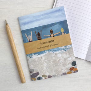 May include: A lined notebook with a cover illustration of four people swimming in the ocean, featuring a tan paper band printed with the text "jamesellis lined notebook | 36 pages". A light wood pen is positioned to the left of the notebook.