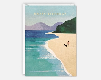 Op de afbeelding: Een wenskaart met een strandscène. De illustratie toont een persoon en een hond op een zandstrand, met de zee en bergen op de achtergrond. De woorden "HAPPY BIRTHDAY" zijn in goud gedrukt.