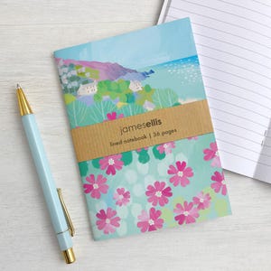 Op de afbeelding: Een gelinieerd notitieboek met 36 pagina's met een illustratie van een kustlandschap op de bovenste helft en roze bloemen op een lichtblauwe achtergrond op de onderste helft, omwikkeld met een bruine papieren band met jamesellis, naast een lichtblauwe pen.