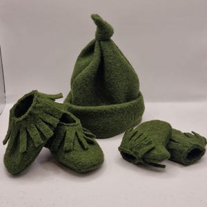 May include: A set of green baby booties, a hat, and mittens. The booties have fringe detailing. The hat has a knotted top.