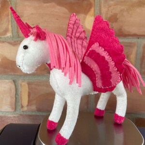 May include: A white felt unicorn with pink accents, including a horn, mane, wings, and hooves. The wings have a layered design. The unicorn is standing on a metallic surface, with a brick background.