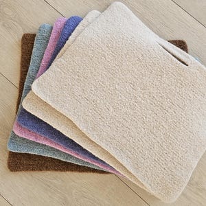 May include: A stack of six textured, rectangular rugs in various colors: brown, teal, pink, purple, and beige. The rugs are made of a soft, plush material and are arranged on a light wood floor.