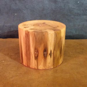 Könnte beinhalten: Ein zylindrischer Holzblock mit flacher Ober- und Unterseite. Die Holzmaserung ist sichtbar, mit vertikalen Streifen aus hellem und dunklem Braun. Der Block ist etwa 15 cm hoch und 15 cm im Durchmesser. Der Hintergrund ist dunkelgrau.