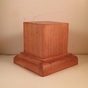 Peut inclure: Un piédestal en bois avec un dessus carré et une base décorative. Le piédestal est en bois brun clair et a une finition lisse.