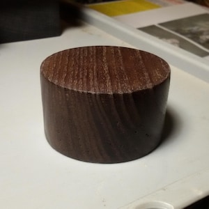 Könnte beinhalten: Ein zylindrisches Holzobjekt mit flacher Ober- und Unterseite. Die Holzmaserung ist auf der Oberseite sichtbar. Das Objekt ist dunkelbraun und liegt auf einer weißen Oberfläche.