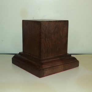 Könnte beinhalten: Ein dunkelbraunes Holzpodest mit quadratischer Oberseite und einem dekorativen Sockel. Das Podest ist aus massivem Holz gefertigt und hat eine glatte Oberfläche.