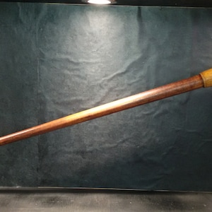 Può includere: Un bastone da passeggio lungo in legno con finitura marrone e gialla. Il bastone ha un anello di metallo vicino alla parte superiore.