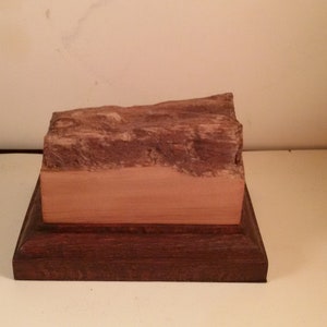 May include: A wooden display stand with a natural wood base and a top layer of reddish-brown wood.
