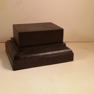 May include: A two-tiered black wooden display stand with a square top and a slightly rounded base.