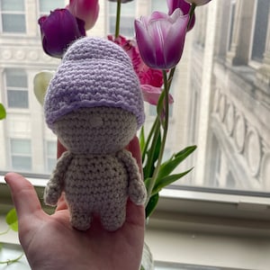 May include: A crocheted amigurumi doll wearing a lavender hat. The doll is standing on a hand with a blurry background of purple tulips.