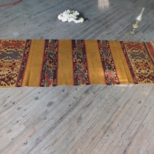 May include: A long, rectangular rug with a geometric pattern in shades of brown, red, blue, and yellow. The rug has a fringe on both ends.