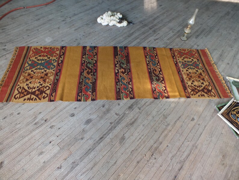 May include: A long, rectangular rug with a geometric pattern in shades of brown, red, blue, and yellow. The rug has a fringe on both ends.