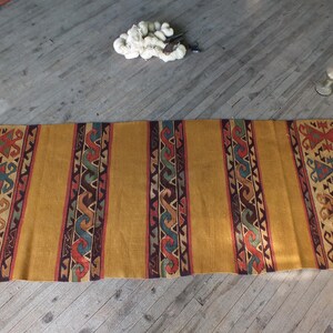 May include: A long, narrow, woven rug with a geometric pattern in shades of brown, red, blue, and green. The rug has a gold background and is laid out on a wooden floor.