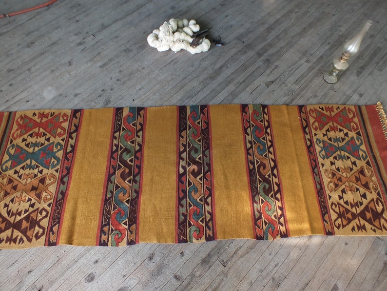 May include: A long, narrow, woven rug with a geometric pattern in shades of brown, red, blue, and green. The rug has a gold background and is laid out on a wooden floor.
