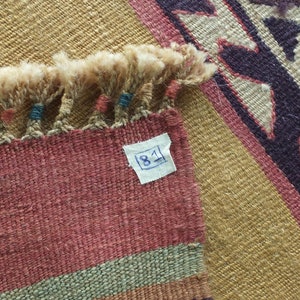 May include: Close-up of a woven rug with a fringe border. The rug has a geometric pattern in shades of brown, beige, and purple. A white tag with the number "81" is attached to the rug.