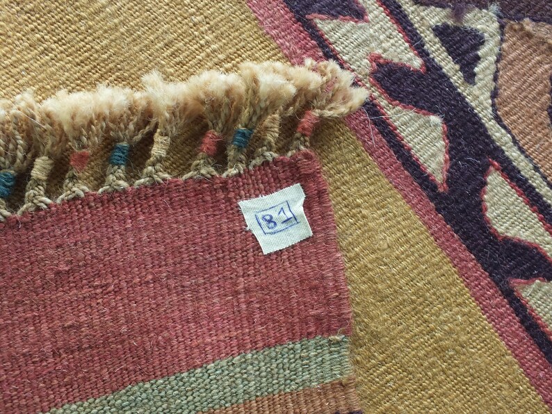 May include: Close-up of a woven rug with a fringe border. The rug has a geometric pattern in shades of brown, beige, and purple. A white tag with the number "81" is attached to the rug.