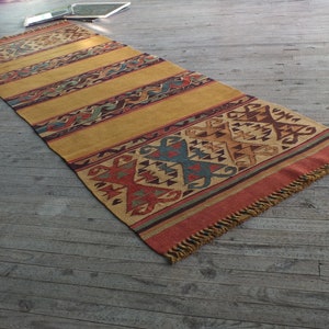 May include: A long, rectangular rug with a geometric pattern in shades of red, orange, yellow, blue, and brown. The rug has a fringe border.