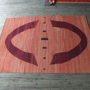 May include: A large, rectangular rug with a reddish-orange background and a dark burgundy design. The design features two curved lines that resemble arches, with a vertical line of dark dots down the center.
