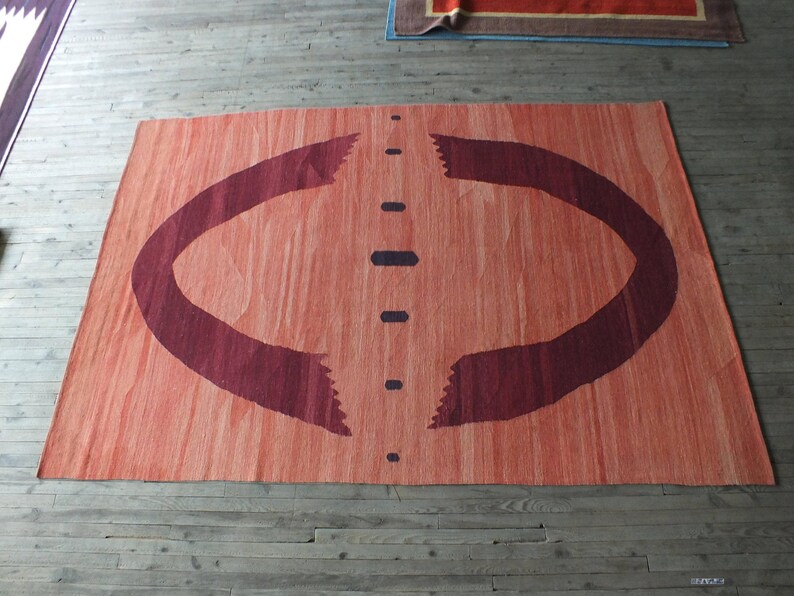 May include: A large, rectangular rug with a reddish-orange background and a dark burgundy design. The design features two curved lines that resemble arches, with a vertical line of dark dots down the center.
