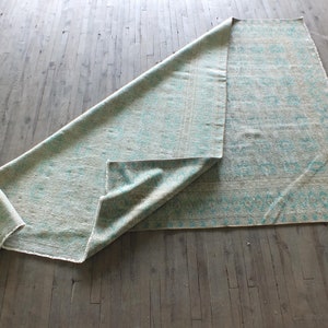 K&ouml;nnte beinhalten: Ein hellblauer und wei&szlig; gemusterter Teppich liegt gefaltet auf einem Holzboden. Der Teppich hat ein geometrisches Muster mit einem hellblauen Rand.