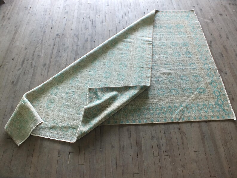 K&ouml;nnte beinhalten: Ein hellblauer und wei&szlig; gemusterter Teppich liegt gefaltet auf einem Holzboden. Der Teppich hat ein geometrisches Muster mit einem hellblauen Rand.
