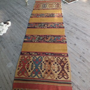 May include: A long, narrow, woven rug with a gold background and colorful geometric patterns. The rug has a fringe on both ends.