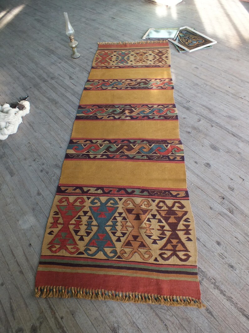 May include: A long, narrow, woven rug with a gold background and colorful geometric patterns. The rug has a fringe on both ends.