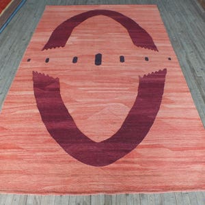 May include: A large, rectangular rug with a peach-colored background and a large, dark purple, abstract design in the center. The design resembles a stylized eye or a pair of wings.