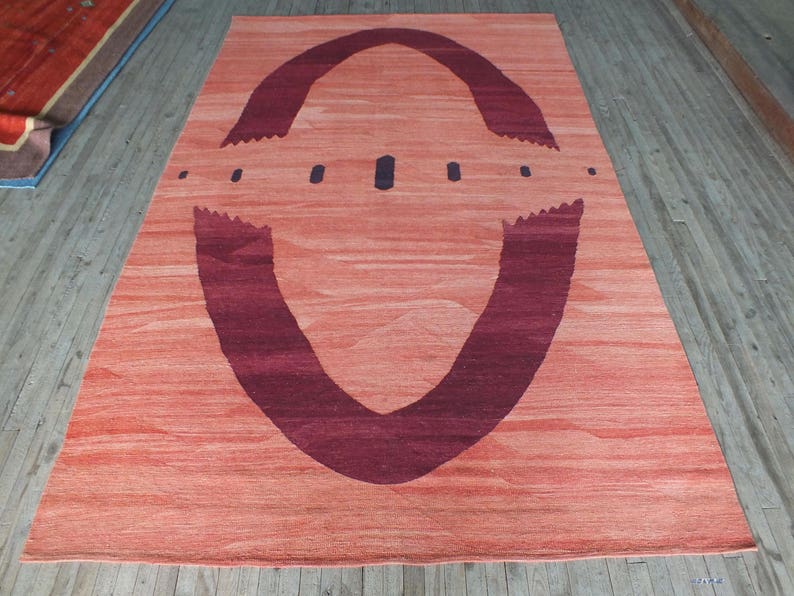 May include: A large, rectangular rug with a peach-colored background and a large, dark purple, abstract design in the center. The design resembles a stylized eye or a pair of wings.