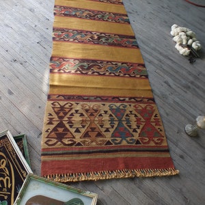 May include: A long, rectangular rug with a traditional Turkish design. The rug has a gold background with red, blue, and green geometric patterns. The rug has a fringe on both ends.