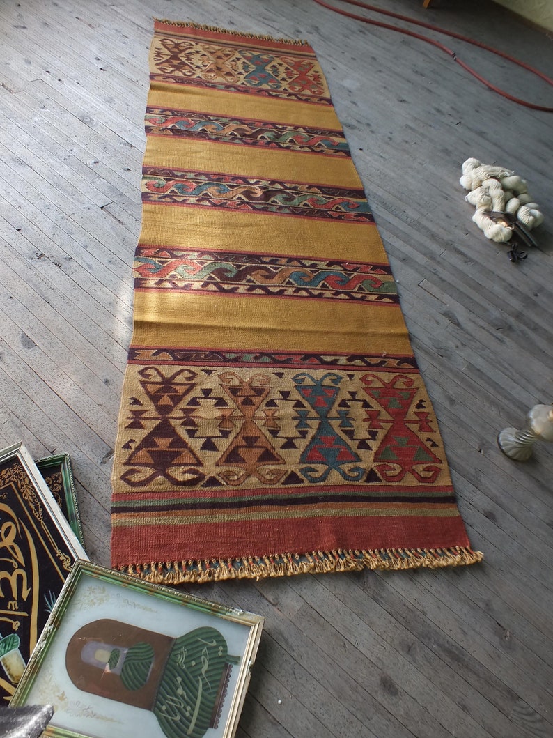 May include: A long, rectangular rug with a traditional Turkish design. The rug has a gold background with red, blue, and green geometric patterns. The rug has a fringe on both ends.