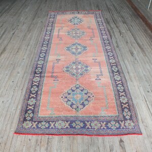 Puede incluir: Una alfombra vintage con fondo color melocotón y un patrón geométrico azul marino, verde y blanco. La alfombra tiene un borde con flecos.