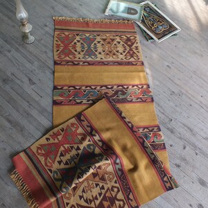 May include: A handwoven rug with a geometric pattern in shades of red, brown, yellow, and blue. The rug has a fringe border and is folded in half.