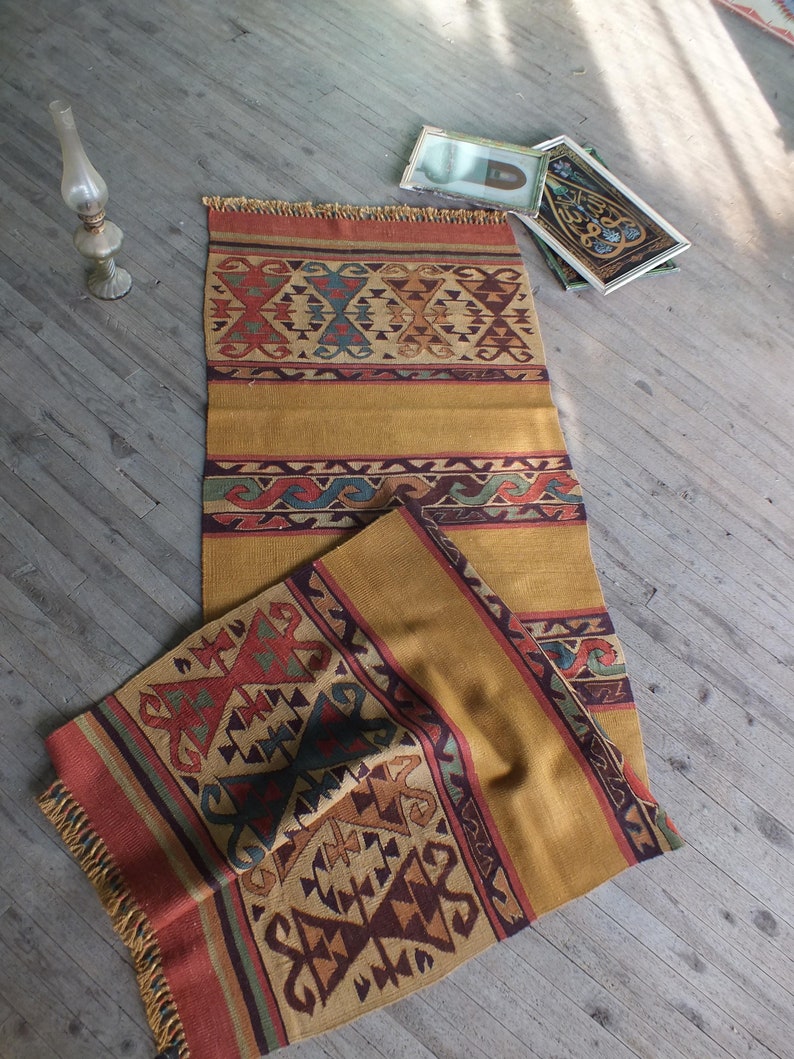 May include: A handwoven rug with a geometric pattern in shades of red, brown, yellow, and blue. The rug has a fringe border and is folded in half.